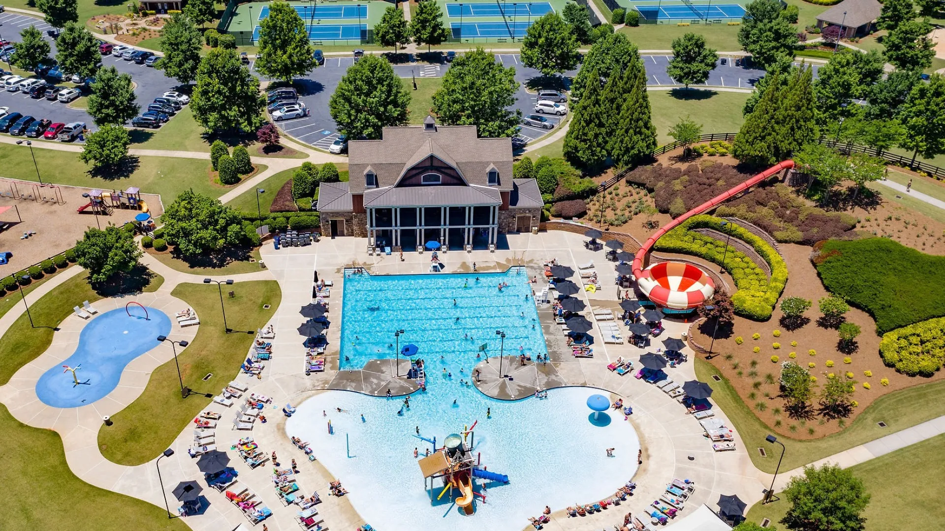 Aerial view of a vibrant community pool area with water slides, playgrounds, tennis courts, and surrounding greenery.